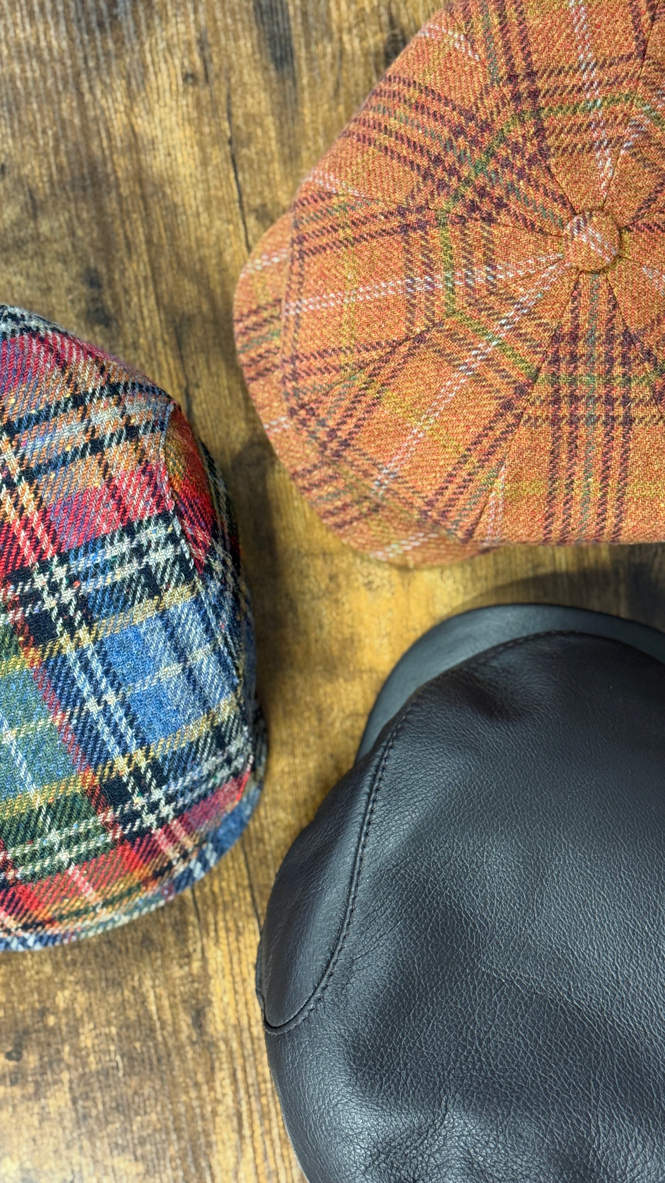 Three different types of caps on a wooden surface: plaid harris Tweed, checkered wool, and dark brown leather.
