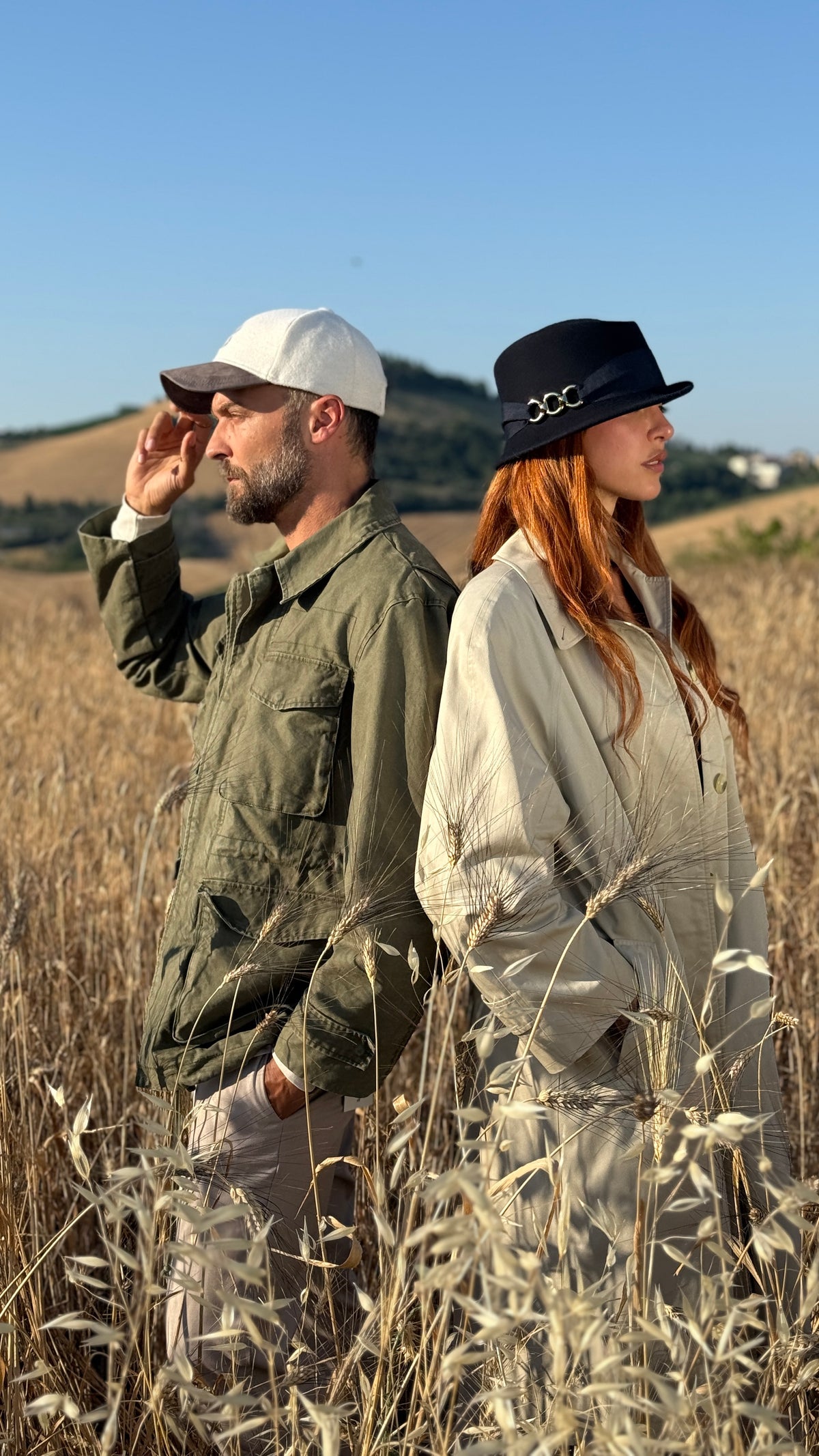 A man and a woman standing back-to-back in a dry grass field under a clear blue sky. The man wears an olive green jacket, beige pants, and a white cap, while the woman wears a light beige jacket and a black bucket hat with a silver logo.