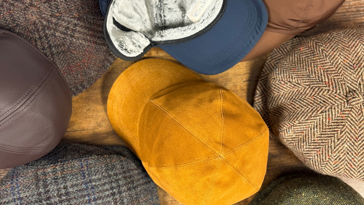 A mustard yellow suede baseball cap is arranged on a wooden surface, surrounded by other textured caps and tweed fabrics in brown, navy, and herringbone patterns.