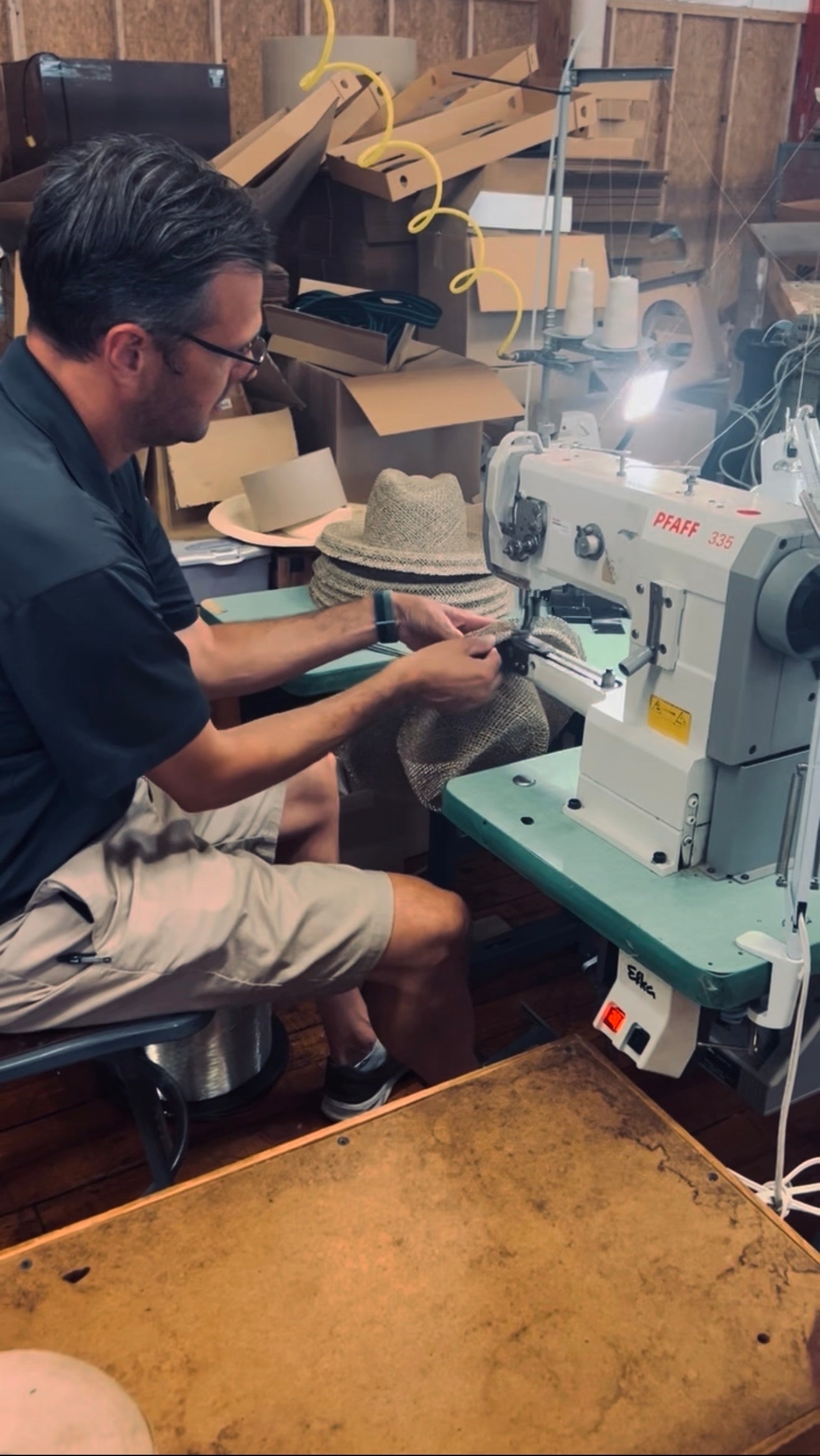 Man working with a sewing machine and hats in a workshop setting
